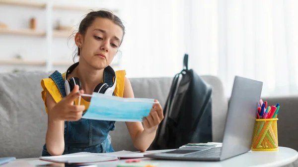 Menina triste segurando máscara médica estudando em casa com laptop — Fotografia de Stock