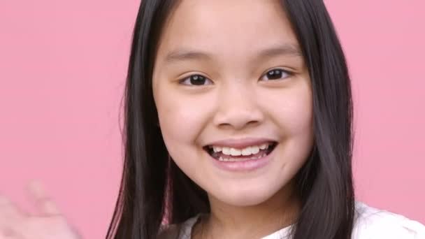 Glada hälsningar. Positiv liten asiatisk flicka vinka hand hej och ler mot kameran, rosa studio bakgrund, närbild — Stockvideo