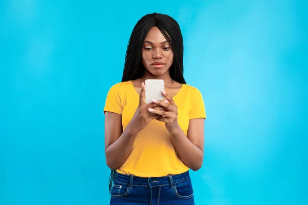 Mujer negra seria usando mensajes de texto de celular de pie sobre fondo azul — Foto de Stock