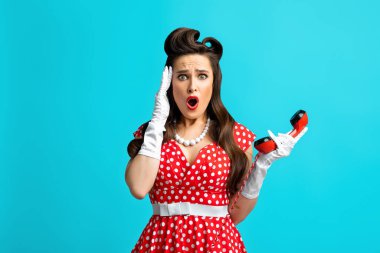 Elinde ev telefonu ahizesi tutan, mavi arka plandaki şakaklarına dokunan, eski moda bir elbise giymiş korkunç bir milenyum kadını.