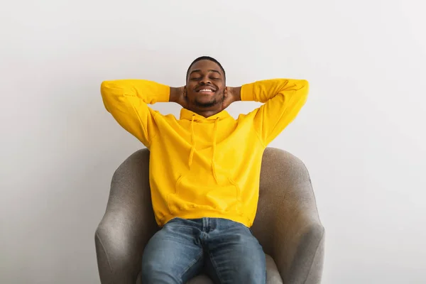 Negro chico sentado con las manos detrás de la cabeza en sillón interior — Foto de Stock