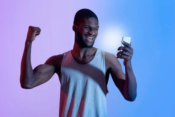 Pria kulit hitam yang bersemangat dengan smartphone membuat gerakan YES, memenangkan taruhan olahraga di lampu neon — Stok Foto