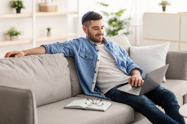 在家里沙发上的笔记本电脑上工作的快乐的男性犹太人 — 图库照片