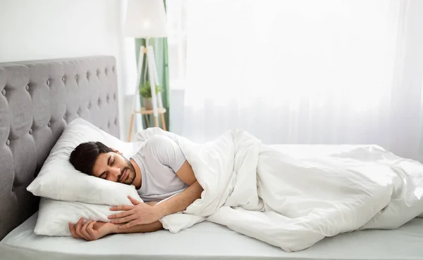 Hora de dormir. cansado milenial árabe hombre dormido, descansando pacíficamente en blanco cómoda cama en el dormitorio, espacio de copia — Foto de Stock
