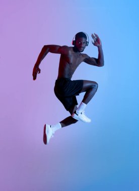 Uzun boylu, formda, kulaklıklı siyahi bir sporcu havada zıplıyor ve neon ışıkta müzik dinliyor.