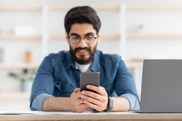 Lächelnder arabischer Freiberufler, der Nachrichten auf dem Smartphone sendet, während er sich am Arbeitsplatz im Home Office entspannt, selektiver Fokus — Stockfoto