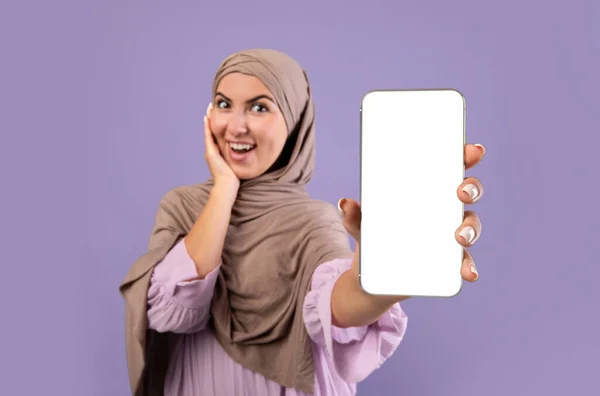 Happy muslim woman in hijab shows cellphone with blank screen for your app or website design, purple background — Stok Foto