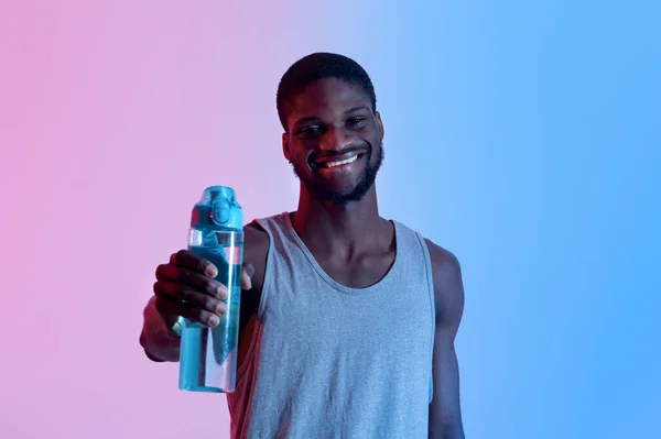 Sonriente joven deportista negro mostrando botella con agua dulce en luz de neón. Fitness, estilo de vida activo, concepto de bienestar —  Fotos de Stock