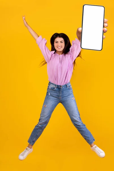 Dama armenia llena de alegría sosteniendo teléfono inteligente, mostrando la pantalla en blanco, saltando sobre el fondo amarillo del estudio —  Fotos de Stock