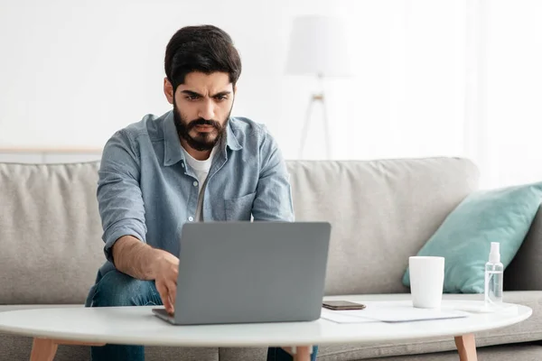 Fernbedienung. Schöner arabischer Typ, der zu Hause im Büro am Laptop arbeitet, auf dem Sofa sitzt und den Computer benutzt, leerer Raum — Stockfoto