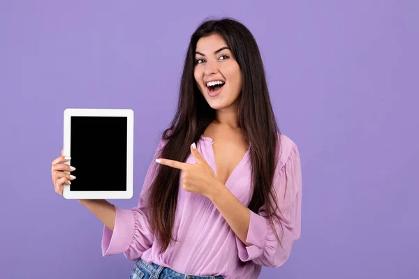 Überprüfen Sie dies. Fröhliche armenische Frau hält und zeigt auf digitale Tablette mit schwarzem leeren Bildschirm, lila Hintergrund — Stockfoto