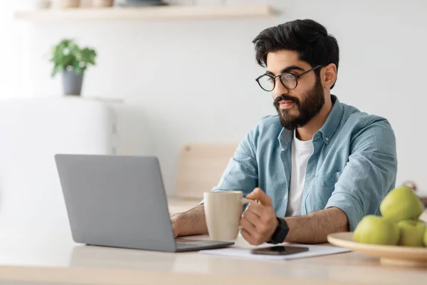 Der moderne Arbeiter in Selbstisolierung. Konzentrierter Araber trinkt Kaffee und arbeitet am Laptop am Tisch in der Küche — Stockfoto