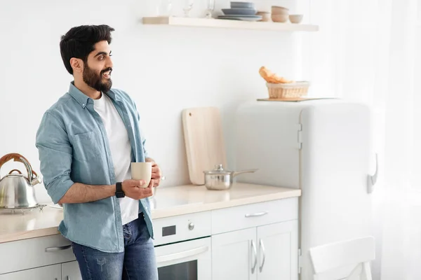 Початок дня. Щасливий арабський чоловік п'є ранкову каву на кухні, стоїть з чашкою гарячого напою, порожній простір — стокове фото