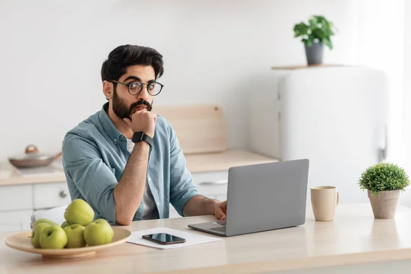 Задумчивый арабский бизнесмен думает о онлайн-проекте, сидя с ноутбуком на кухне и глядя на камеру, свободное пространство — стоковое фото