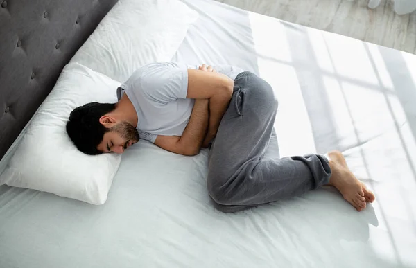 Ból brzucha. młody arabski facet cierpi ból brzucha dotykając ból brzucha leżąc w łóżku, powyżej widok — Zdjęcie stockowe