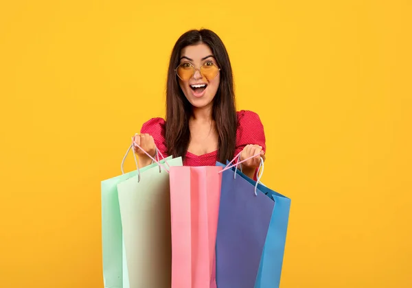 Un cliente feliz. Mujer armenia llena de alegría en gafas de sol sosteniendo bolsas de compras brillantes, posando sobre fondo amarillo —  Fotos de Stock