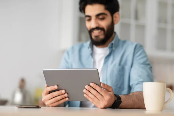 Tiempo libre con dispositivo. Hombre árabe feliz leyendo e-book en tableta digital o navegar por Internet mientras está sentado en la cocina — Foto de Stock