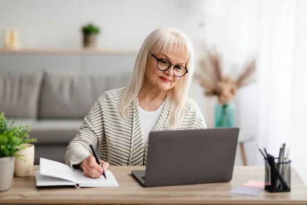 Mature woman writing and using laptop at home — Stock Photo, Image