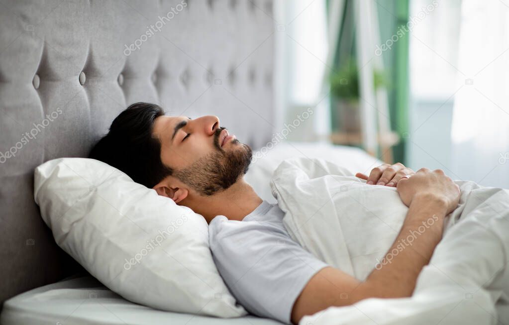 Deep male sleep. Young arab guy sleeping peacefully in his comfortable bed at home, lying with closed eyes on back