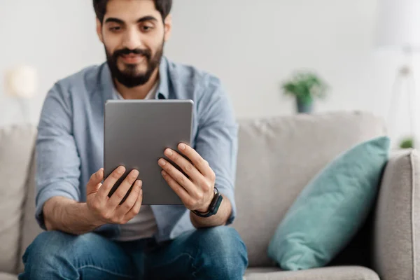 Millennial arab man sitting on sofa with tablet computer, working or study online at home, focus on device — Stock fotografie