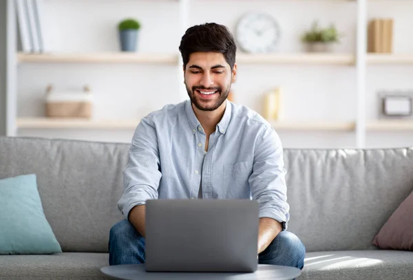 Koncepcja pracy na własny rachunek. Wesoły arabski biznesmen pracujący na laptopie online i uśmiechnięty siedząc na kanapie w domu — Zdjęcie stockowe