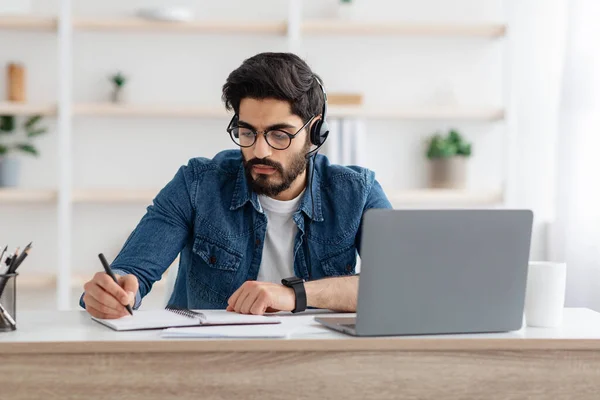 Internetes oktatási koncepció. Arab férfi fülhallgató írás notebook online előadás során a laptop, otthon ül — Stock Fotó