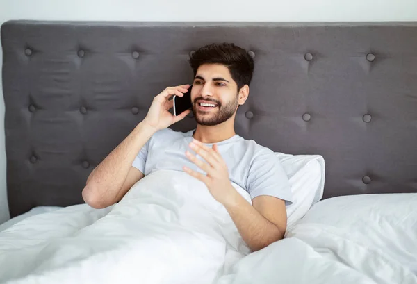 Boldog arab srác beszél okostelefonon, reggel felhív valakit, és mobilbeszélgetést folytat az ágyban — Stock Fotó