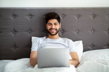 Her zaman online. Mutlu Arap adam uzaktan bilgisayarla çalışıyor, yatakta oturuyor, uyanıyor ve bilgisayar kullanıyor, boşluğu kopyalıyor.