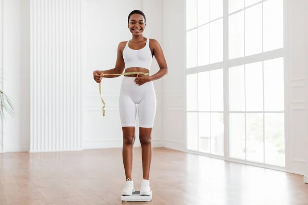 Mujer negra feliz medición de la cintura con cinta de pie en la escala — Foto de Stock