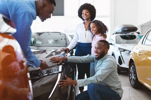 Feliz família negra escolhendo carro novo, vendedor mostrando-lhes automóvel de luxo na loja concessionária de automóveis — Fotografia de Stock