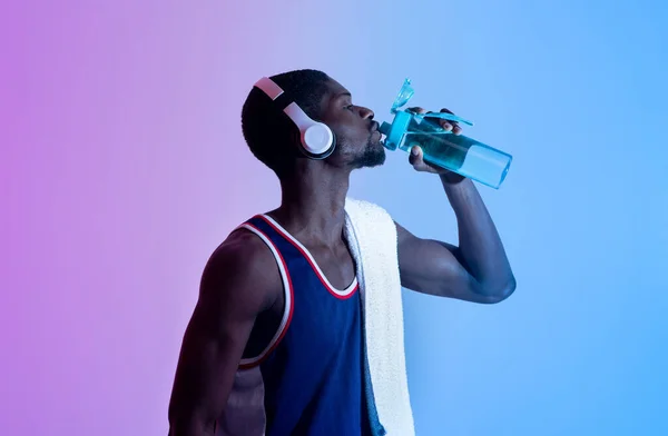 Jovem desportista preto com toalha de banho e fones de ouvido bebendo água de garrafa em luz de néon — Fotografia de Stock