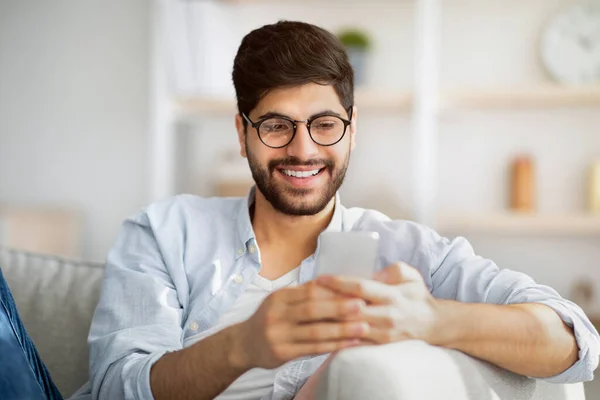 유용 한 앱. 안경을 쓴 긍정적 인 남자가 소파에 앉아서 smatphone 을 사용하거나 인터넷을 보거나 메시지를 보내고 있습니다. — 스톡 사진