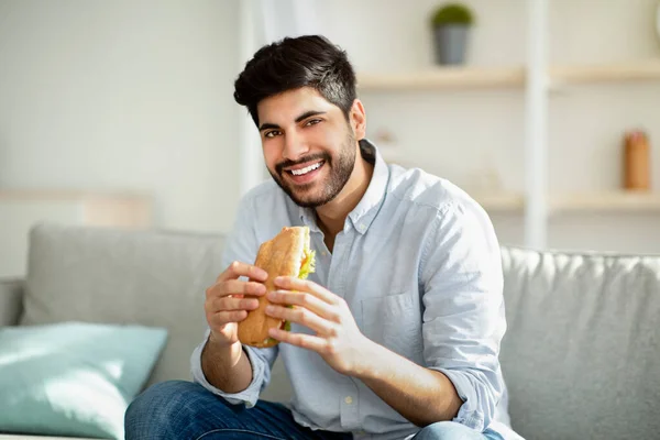 Fuska på måltiden. Glad arabisk man äter god smörgås och ler mot kameran, sitter på soffan i vardagsrummet hemma — Stockfoto