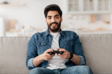 Rahat ortadoğulu adam evde joystick ile video oyunu oynuyor, kanepede oturuyor, yer kopyalıyor.
