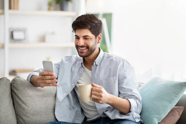 Concepto de vida en línea. Hombre árabe positivo sosteniendo teléfono inteligente moderno y taza de café en casa, espacio libre — Foto de Stock