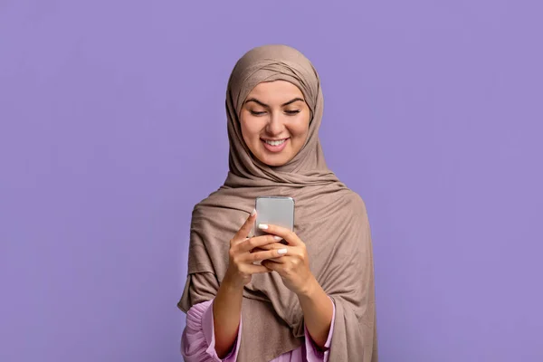 Aplikasi keren. Wanita muslim muda yang bersemangat dalam jilbab menggunakan smartphone, SMS atau browsing internet, latar belakang ungu — Stok Foto
