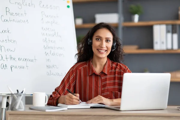 Female teacher in headphones sitting at desk looking at camera