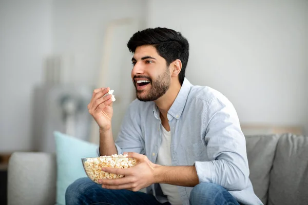 Домашние развлечения. Возбужденный араб ест попкорн и смотрит телевизор, проводит свободное время дома, свободное место — стоковое фото
