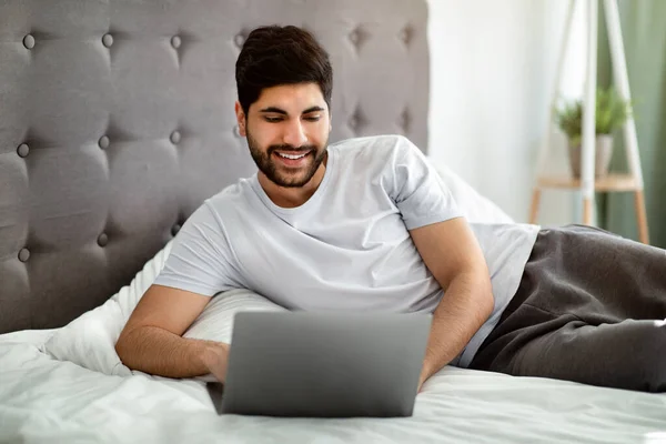 Praca online, nauka, kursy, nowa normalna i bądź bezpieczny podczas COVID-19. Szczęśliwy arabczyk leżący na łóżku, patrzący na laptopa — Zdjęcie stockowe