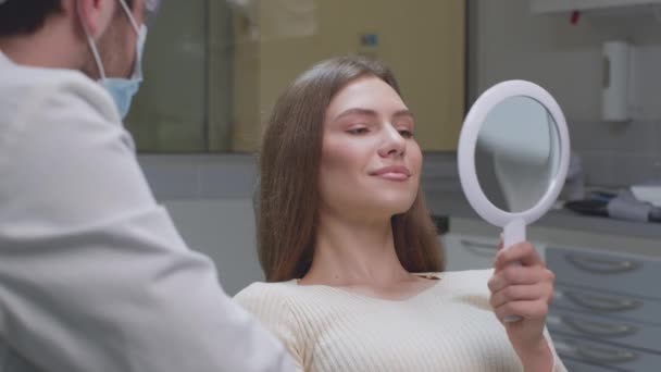 Mladá pozitivní žena se dívá na zrcadlo po ošetření zubů postupy, těší výsledek a usmívá se k lékaři — Stock video