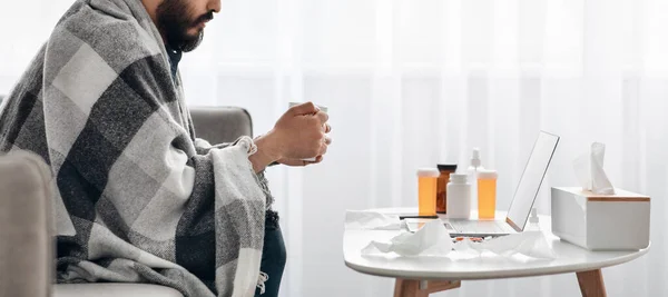 Concepto de fiebre. Enfermo sensación de malestar y beber té caliente, sentado en el sofá y viendo la película en el ordenador portátil, vista lateral, cosecha —  Fotos de Stock