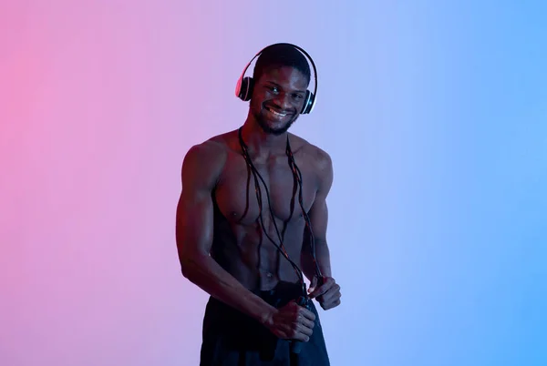 Beste Playlist für Ausdauertraining. Cooler lächelnder schwarzer Sportler mit Kopfhörern und Springseil posiert im Neonlicht — Stockfoto