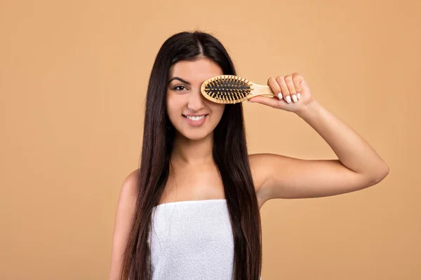 Concept de soins capillaires. Joyeux arménien jeune femme avec de longs cheveux clairs couvrant oeil avec brosse à cheveux, fond beige — Photo