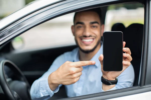 Uśmiechnięty arabski biznesmen siedzi w samochodzie, pokazuje smartfona, makietę — Zdjęcie stockowe