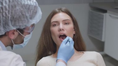 Genç bir kadın dişçi kliniğinde oturuyor, profesyonel dişçi üniforması giyiyor ofiste aynayla dişlerini inceliyor.