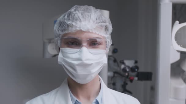 Крупный план портрета профессионального стоматолога в защитной маске и одноразовой шляпе, смотрящего в камеру в офисе — стоковое видео