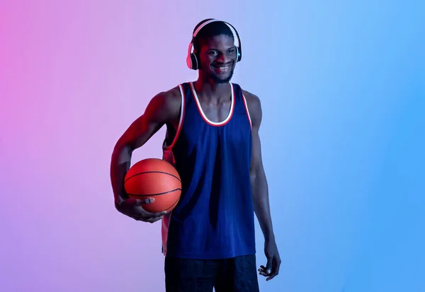 Pemain bola basket hitam yang ceria menikmati musik di headphone, memegang bola dan tersenyum di lampu neon — Stok Foto