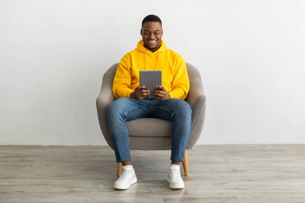 Positivo hombre negro usando tableta sentado cerca de la pared gris en interiores —  Fotos de Stock