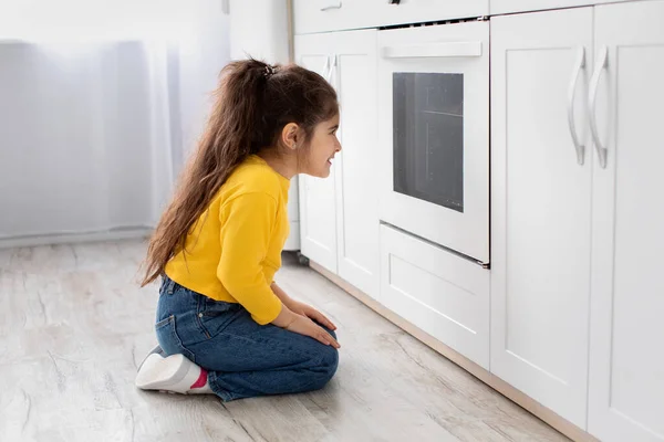 Menina bonito sentado perto do forno na cozinha e olhando para dentro — Fotografia de Stock