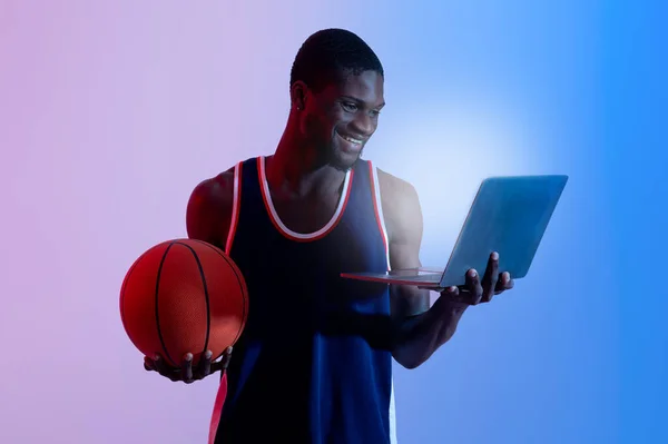 Cheerful African American sports fan holding laptop pc and basketball in neon light — Stok Foto